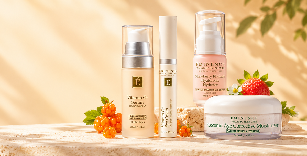 A set of four Eminence Cosmetics skincare products arranged on a textured beige stone surface with decorative strawberries and orange berries. The products include a Vitamin C Serum, a tube, a Strawberry Bladedness Hydrating Treatment, and a Coconut Age Corrective Moisturizer, set against a warm, sun-dappled beige background with soft green leaves.