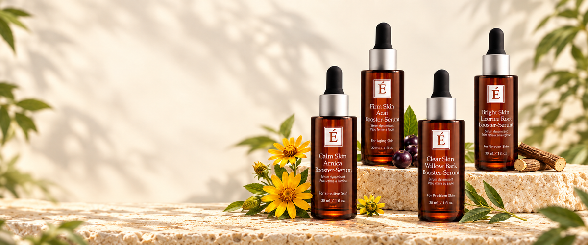 Four amber glass dropper bottles of Elixir Skin botanical serum arranged on a stone surface with yellow flowers and green leaves against a softly lit beige background.