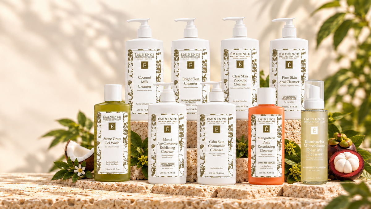 A collection of eminence skincare products arranged on a stone pedestal with tropical props like a coconut, flower, and mangosteen. The products include cleansers, toners, and gels in white, green, and orange packaging with pump and spray dispensers.