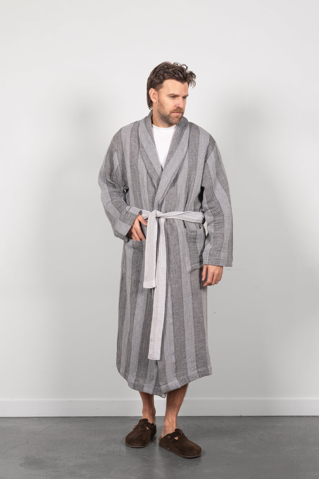 A man wearing a gray vertically striped muslin bathrobe with a white belt, standing against a light gray wall.