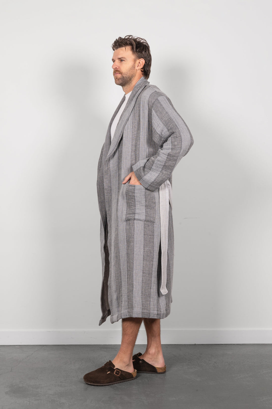 A man wearing a long gray striped muslin bathrobe with a tie belt, standing in profile against a light gray wall and concrete floor.