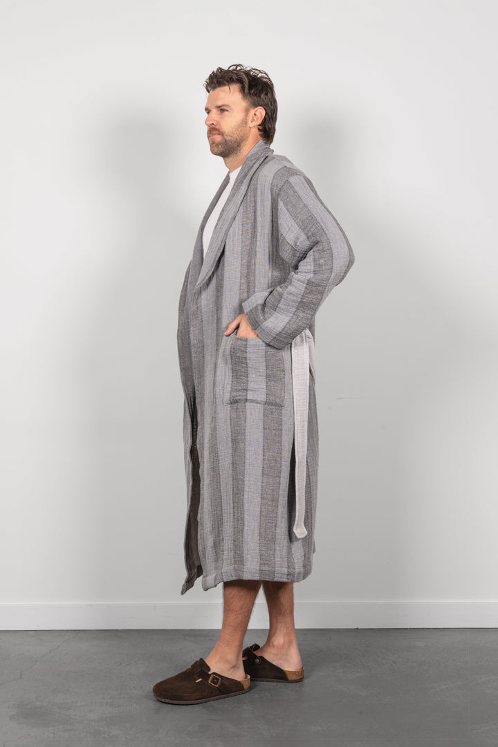 A man wearing a long gray striped muslin bathrobe with a tie belt, standing in profile against a light gray wall and concrete floor.