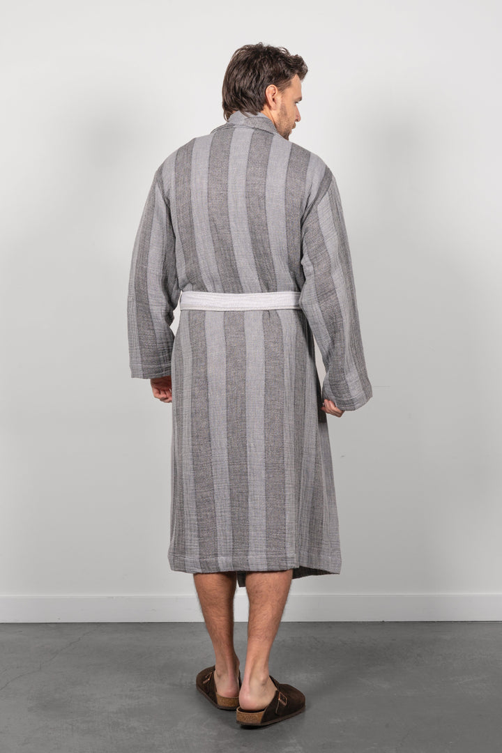 A man viewed from behind wearing a gray vertically striped muslin bathrobe with a white belt, paired with brown leather loafers, standing against a light gray wall.