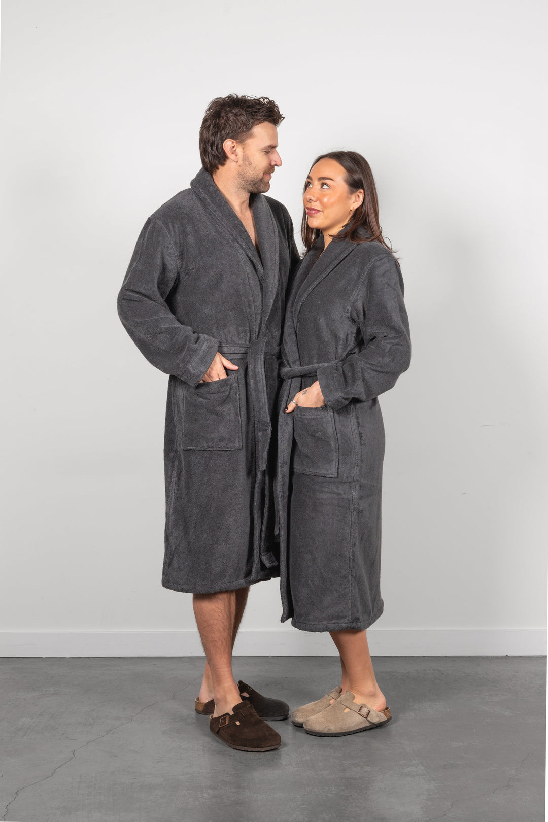A man and a woman standing side by side wearing matching dark gray plush bathrobes and casual slippers, photographed against a plain light gray studio wall and floor.
