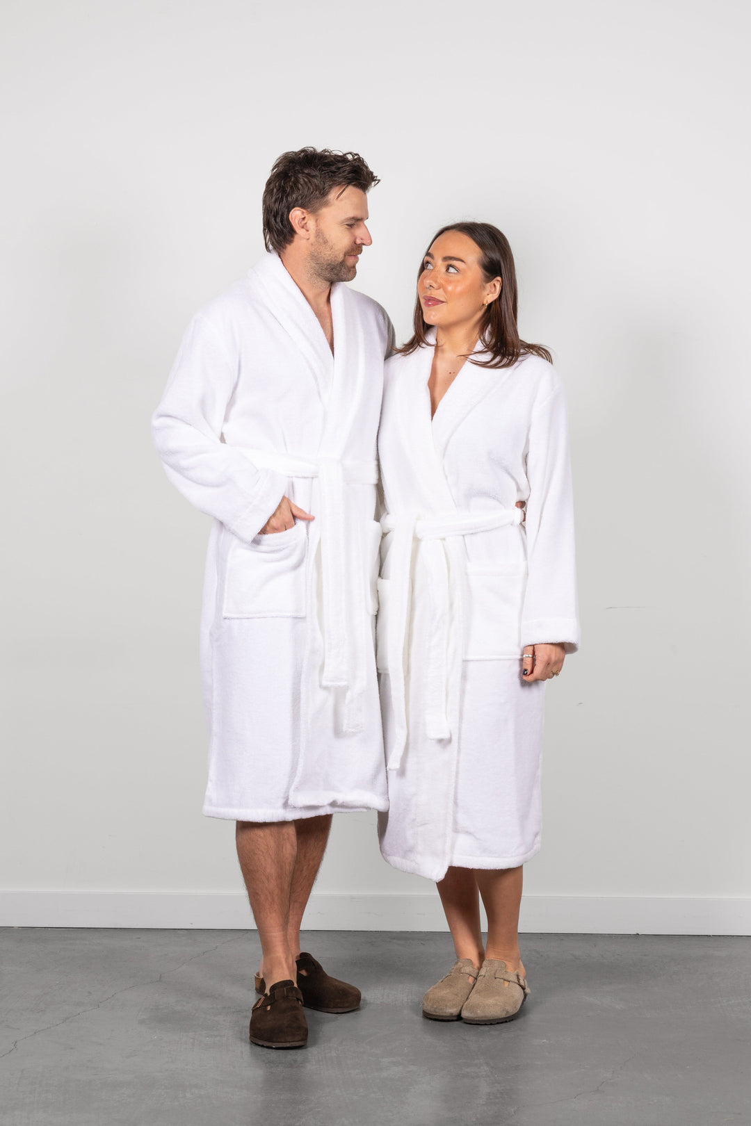A man and a woman standing side by side wearing matching white bathrobes and slippers, photographed against a plain light gray wall and gray floor.