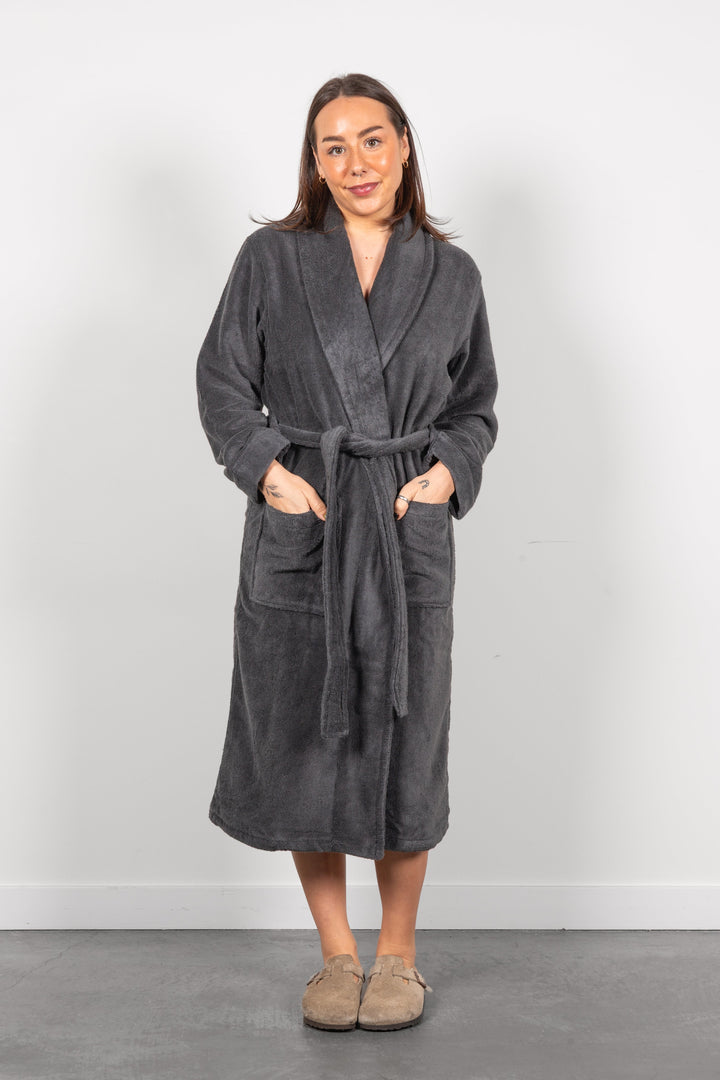 A woman wearing a dark gray plush bathrobe with a tie belt and front pockets, standing against a light gray wall.