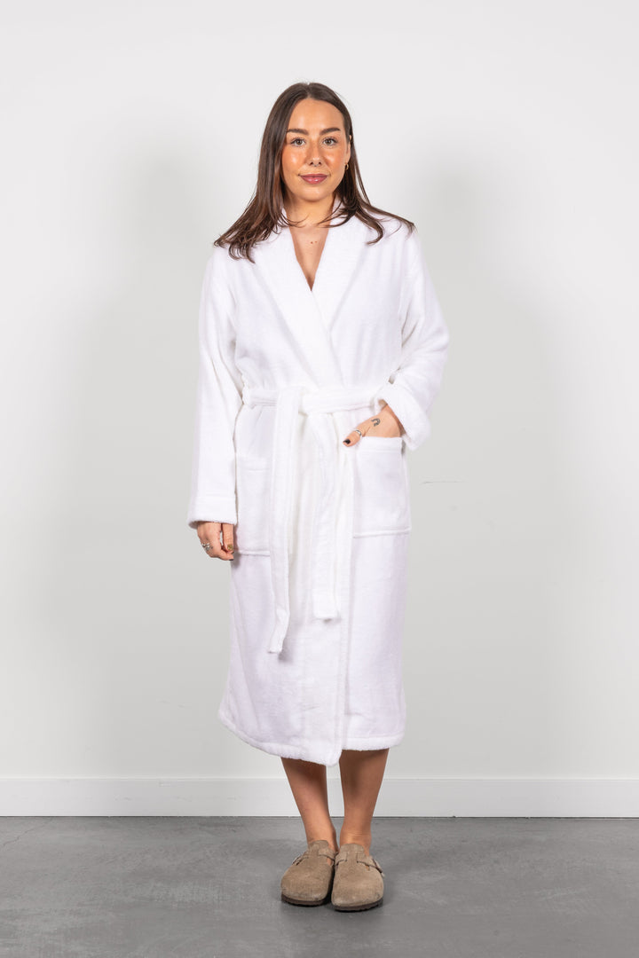 A woman wearing a white terry bathrobe with a tie belt and front pockets, standing against a plain light gray wall on a gray floor.